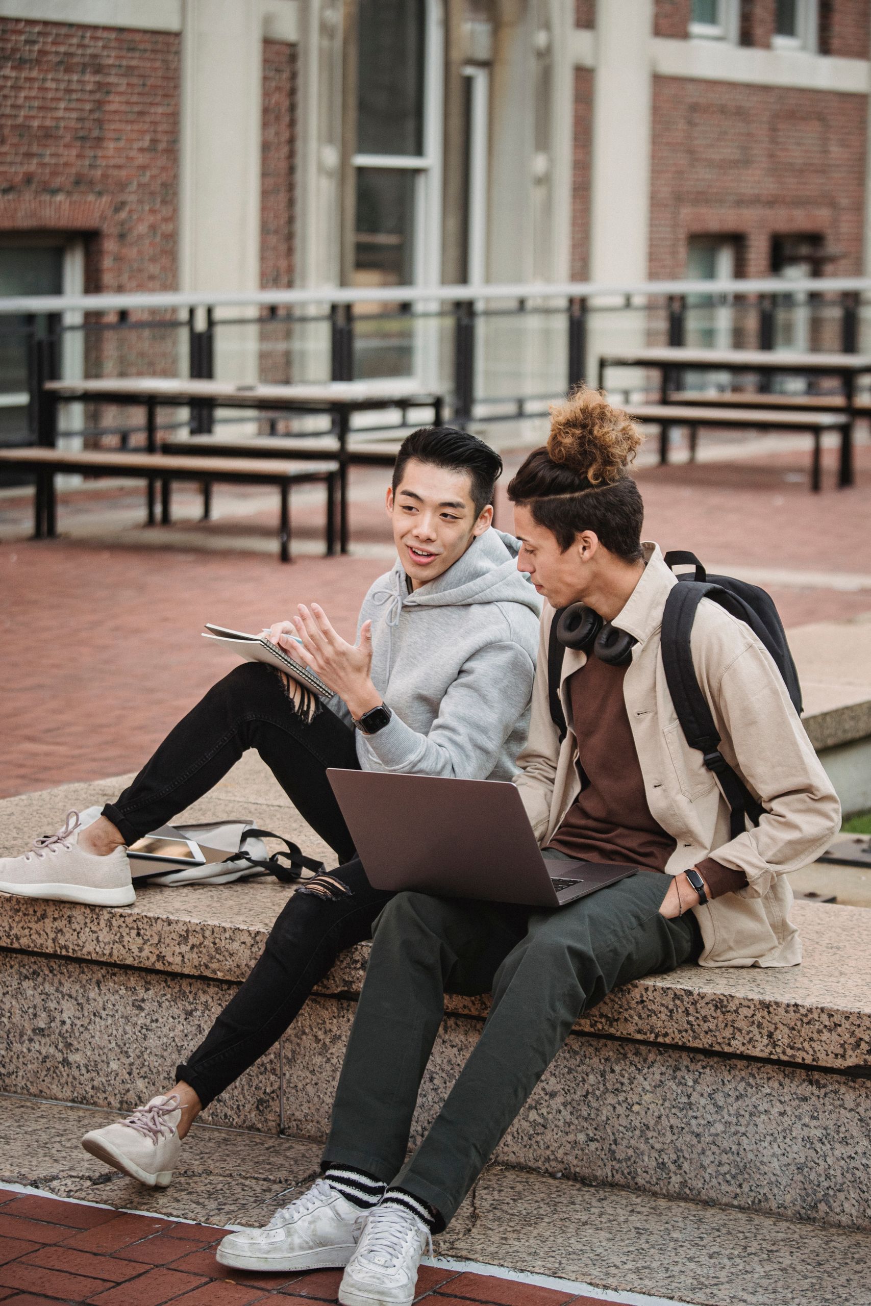 Full length of thoughtful young multiracial male students in casual outfits sitting on stone border with netbook and notepad and discussing project