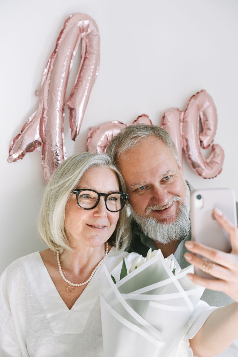Happy senior couple taking a selfie with love-themed balloon backdrop indoors.