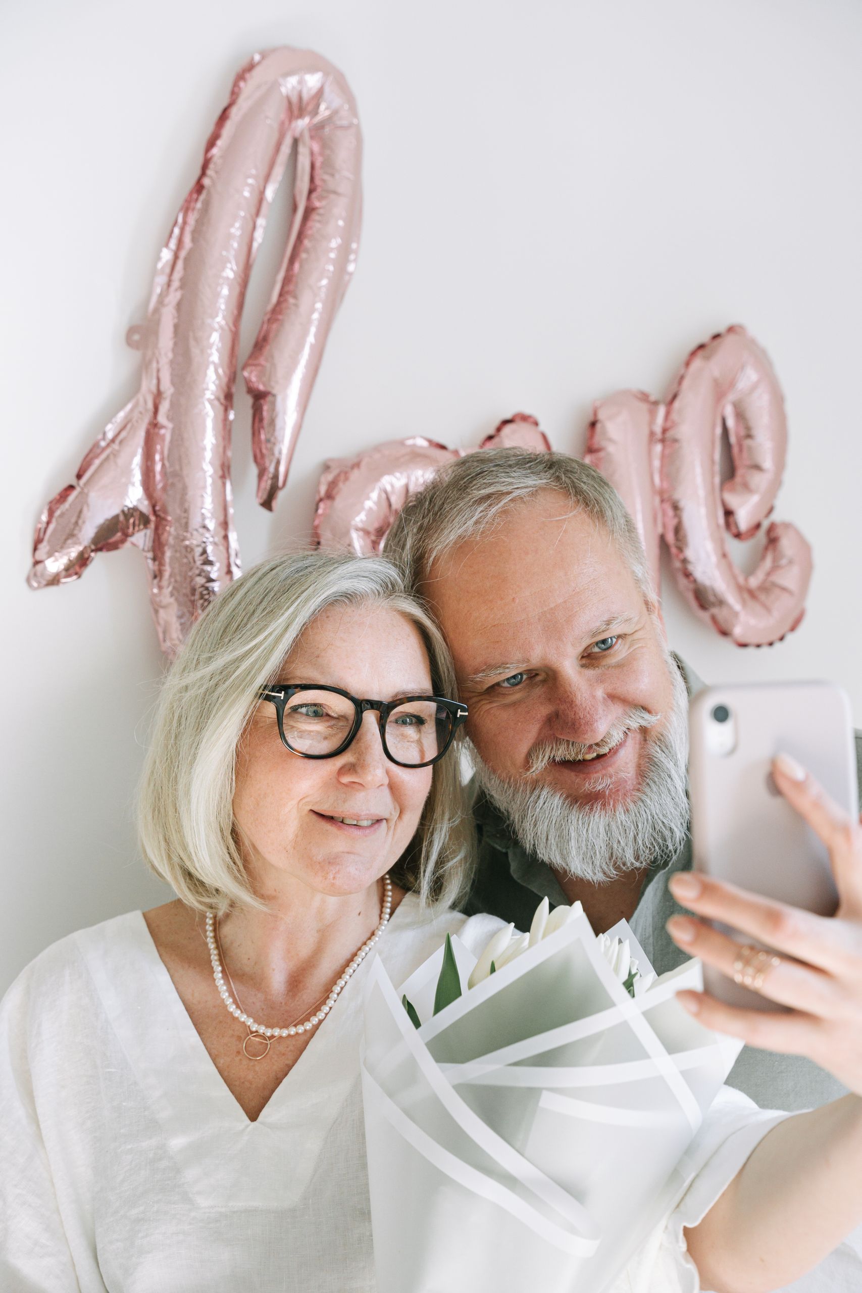 Happy senior couple taking a selfie with love-themed balloon backdrop indoors.