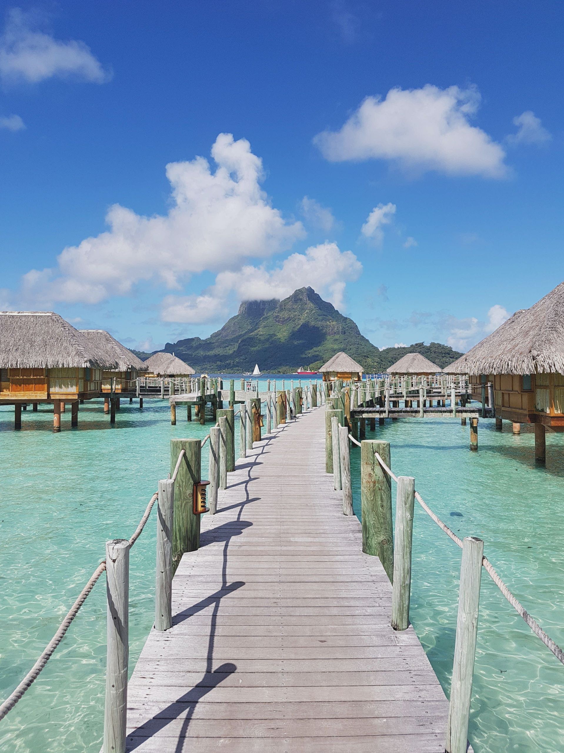 Idyllic tropical scene with wooden walkways and overwater bungalows overlooking a mountain on a sunny day.