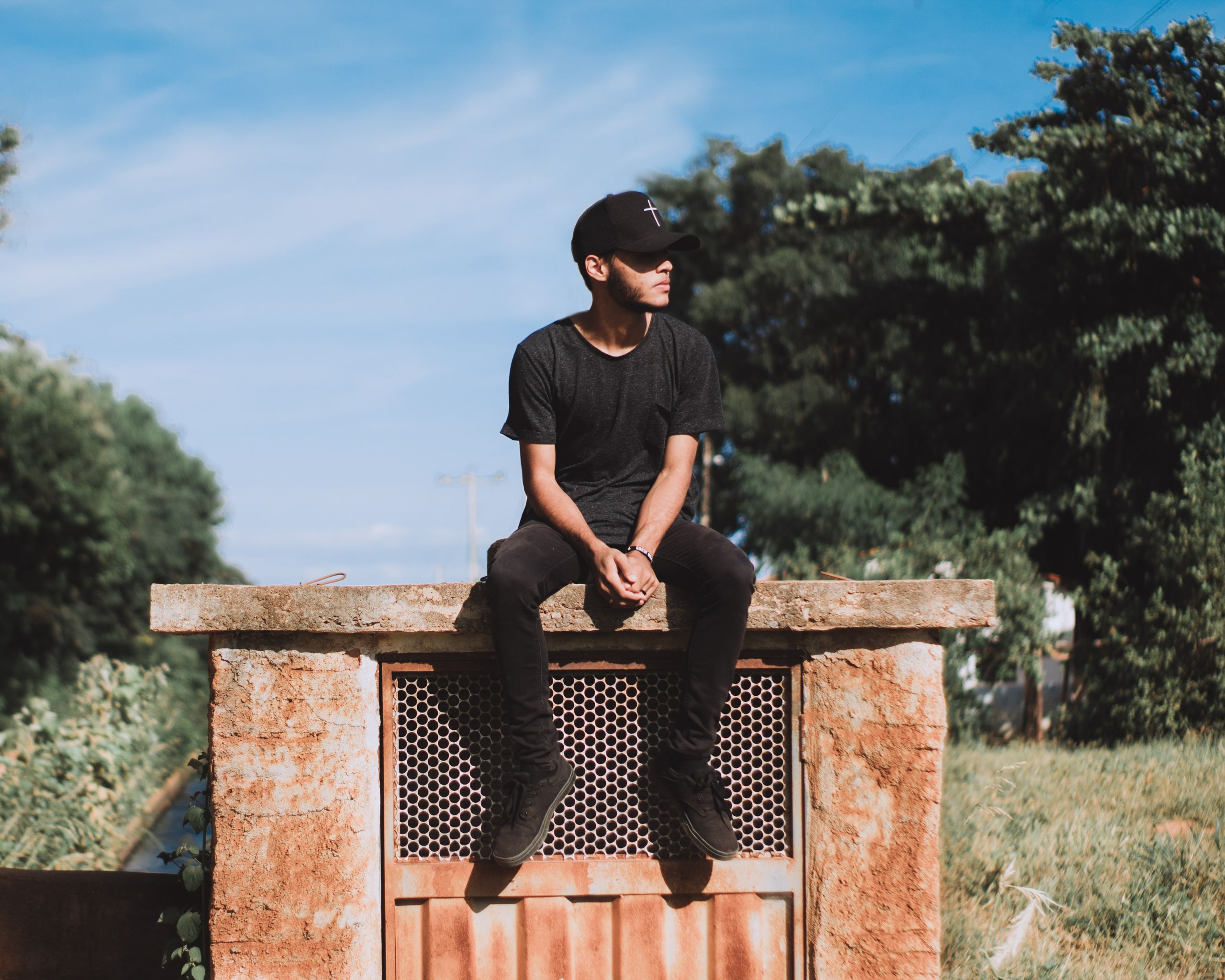 Man in black casual wear sitting on a rustic outdoor structure under the blue sky.