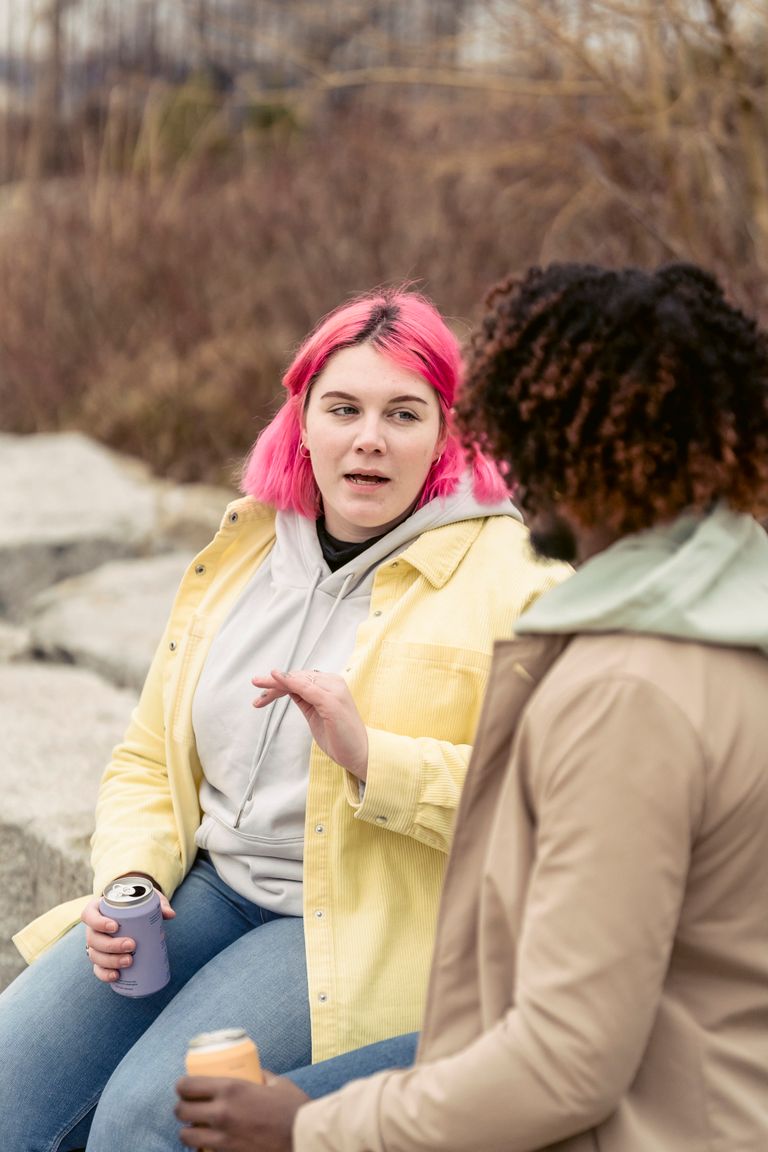 Plump woman with pink hair having conversation with faceless African American man while sitting on stones with cans of soda