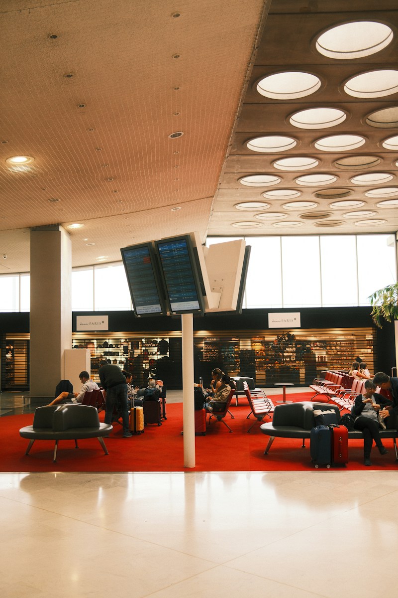 People waiting in an airport terminal with red carpet and flight information screens