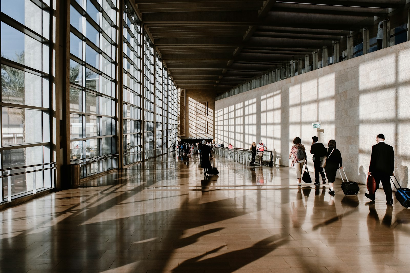 People walking through a sunlit airport terminal with large glass windows and shadows