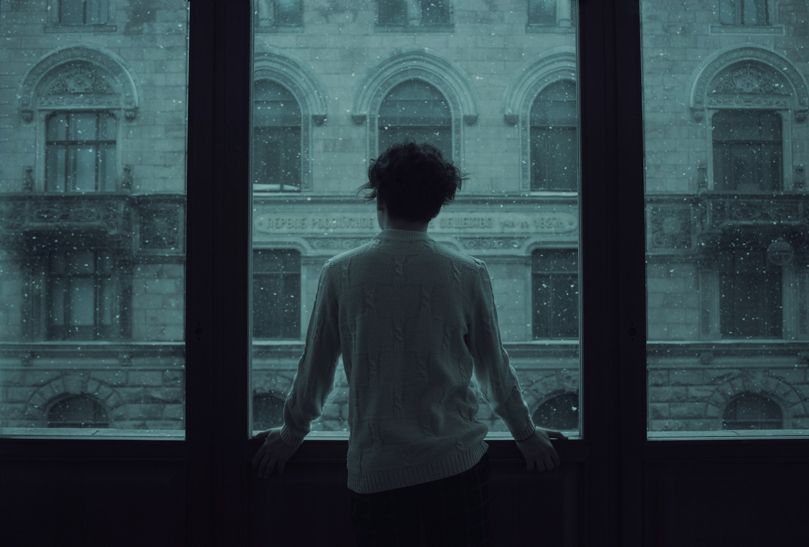 A person in a white sweater looking out a window at falling snow