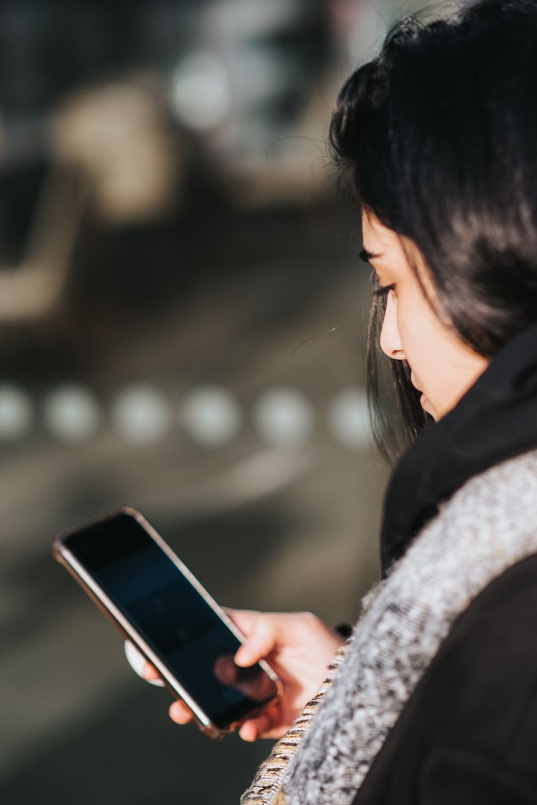 Side view of young ethnic woman in warm clothes and scarf browsing mobile phone on street