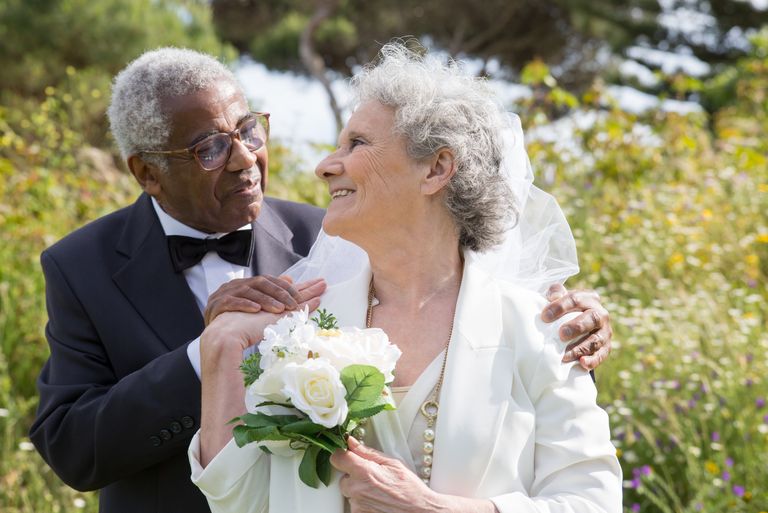 Smiling elderly couple in wedding attire embracing outdoors with flowers.