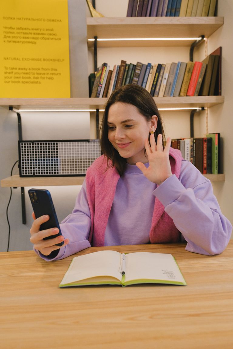 Smiling young woman waving during a video call in a cozy study space with bookshelves.