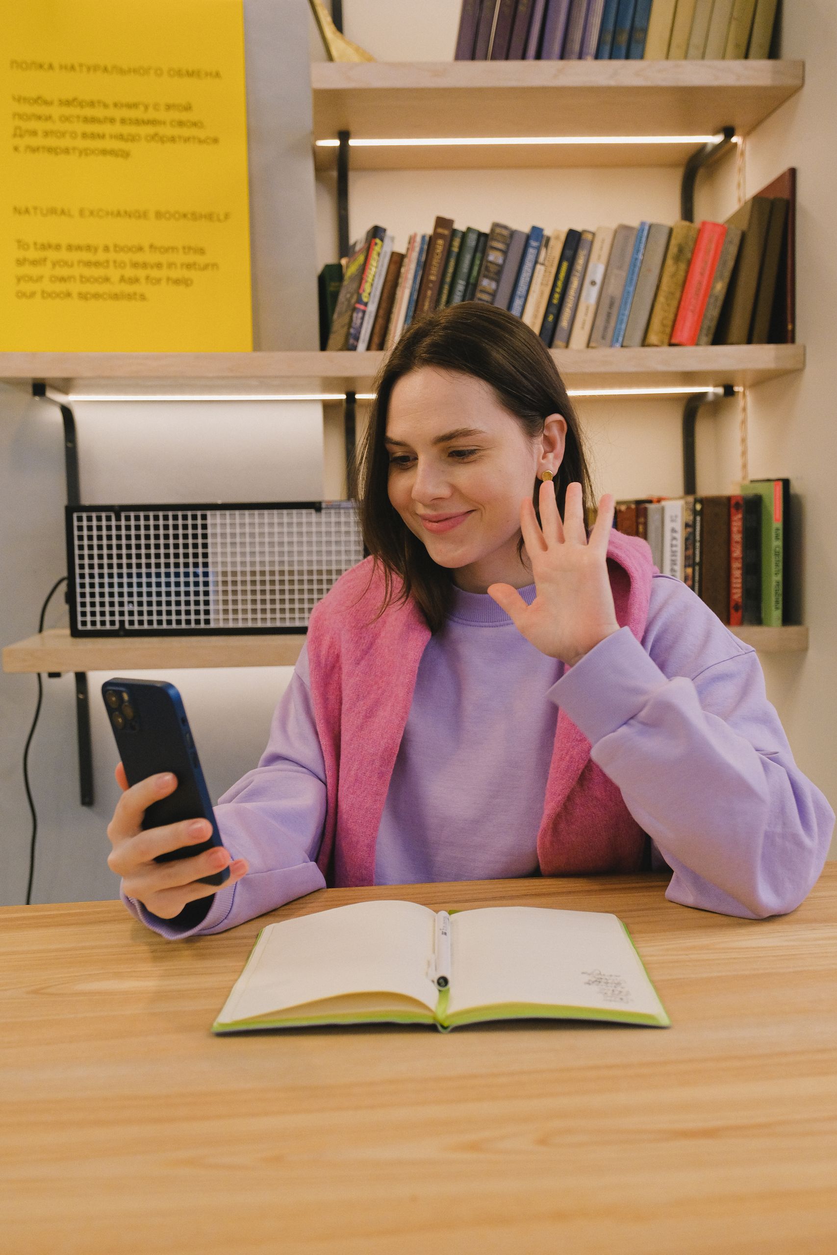 Smiling young woman waving during a video call in a cozy study space with bookshelves.