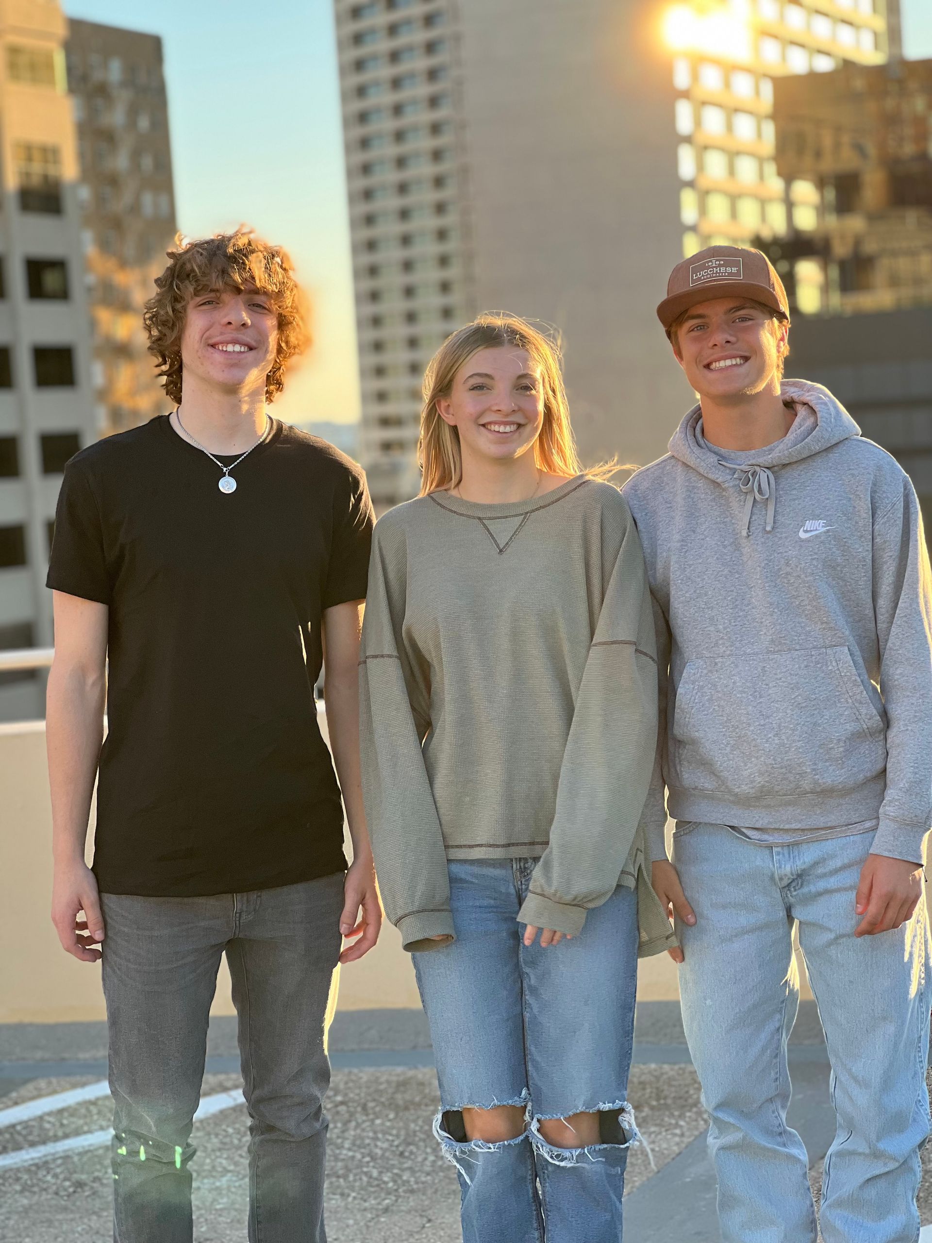 Three teenagers smiling together on a rooftop with city buildings in the sunset background.
