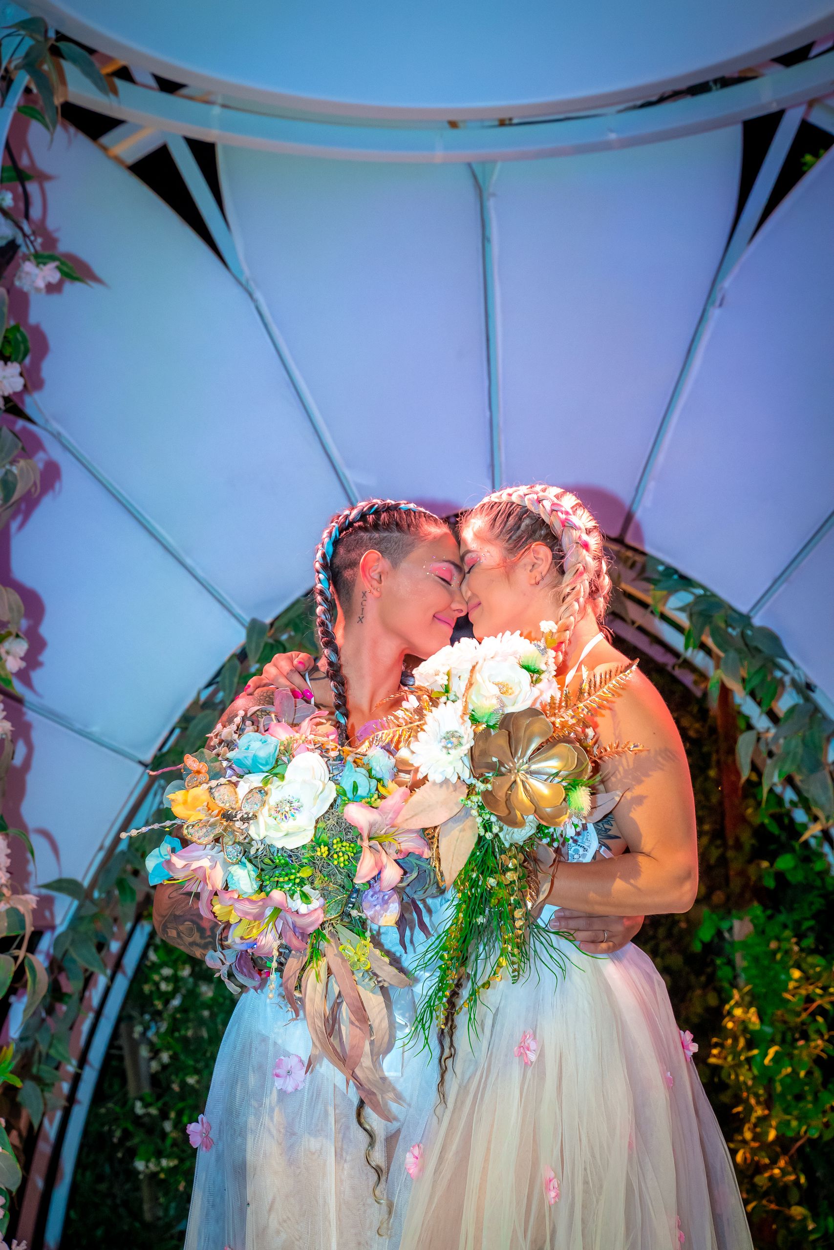 Two brides sharing an intimate moment under a floral arch at a wedding.