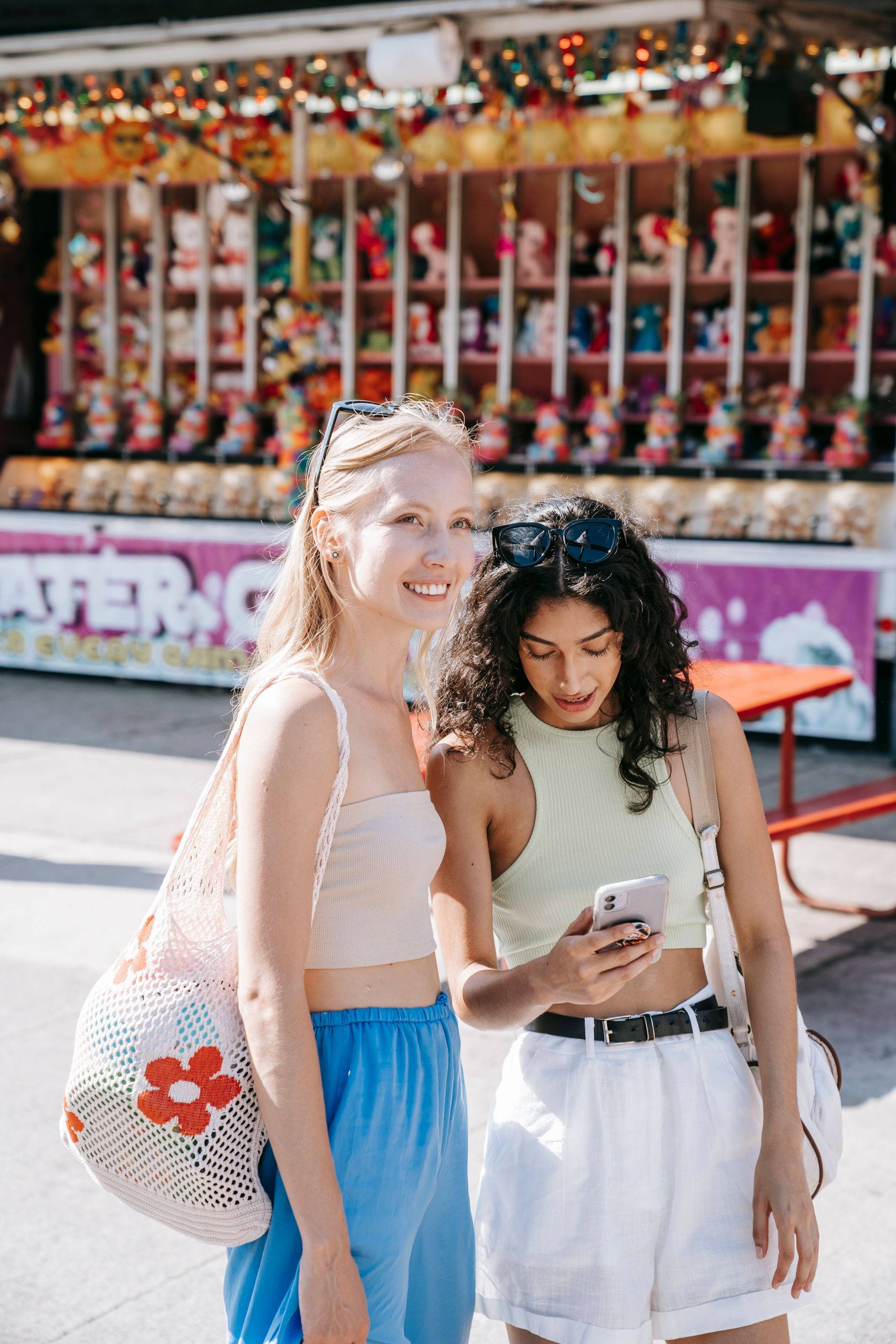 Two women enjoying summer at an outdoor fair, sharing smiles and checking a phone.