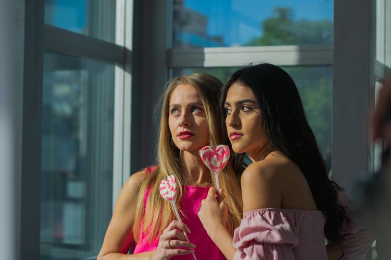 Two young women holding heart-shaped lollipops, bathed in warm sunlight, creating a vibrant and cheerful atmosphere.