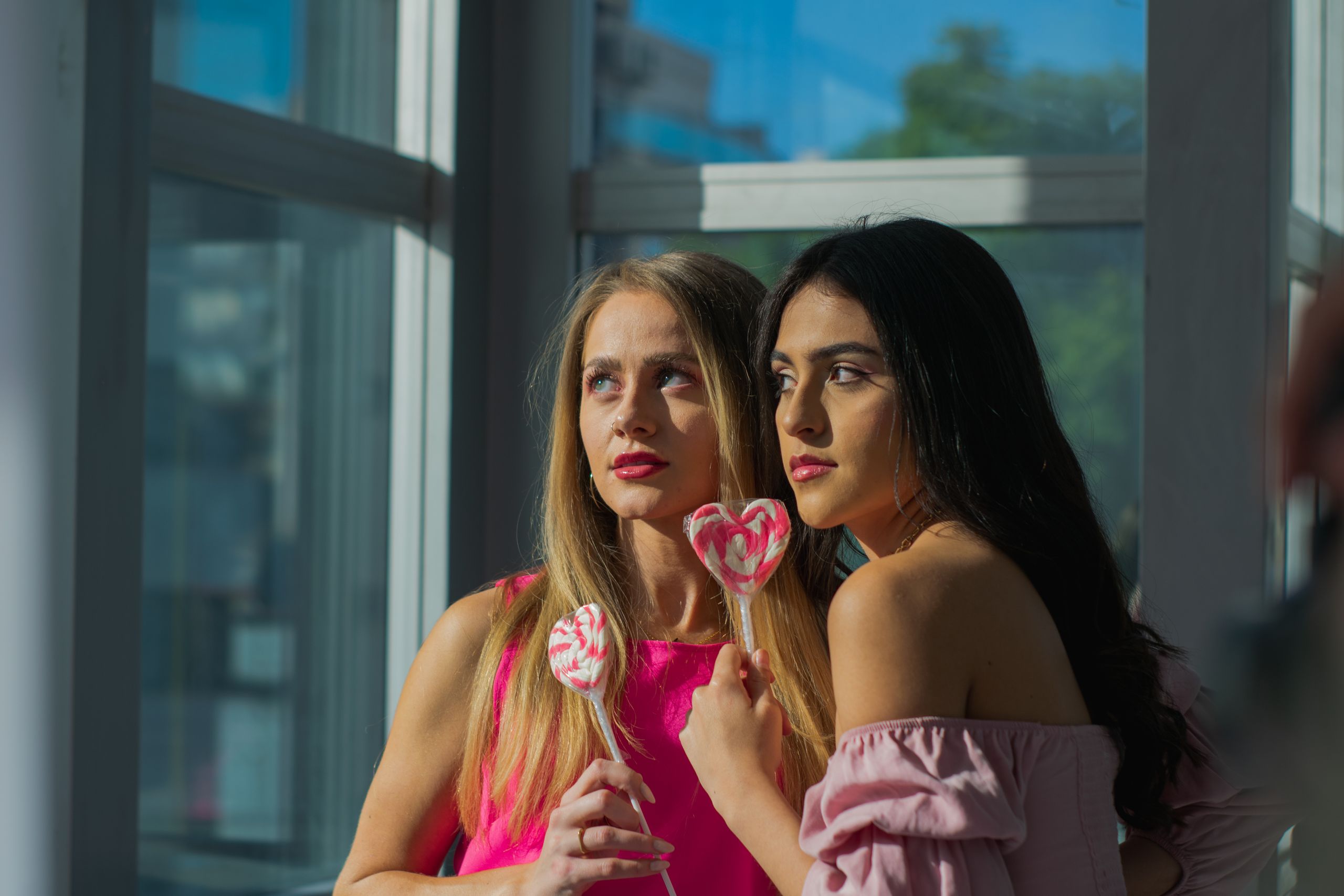 Two young women holding heart-shaped lollipops, bathed in warm sunlight, creating a vibrant and cheerful atmosphere.