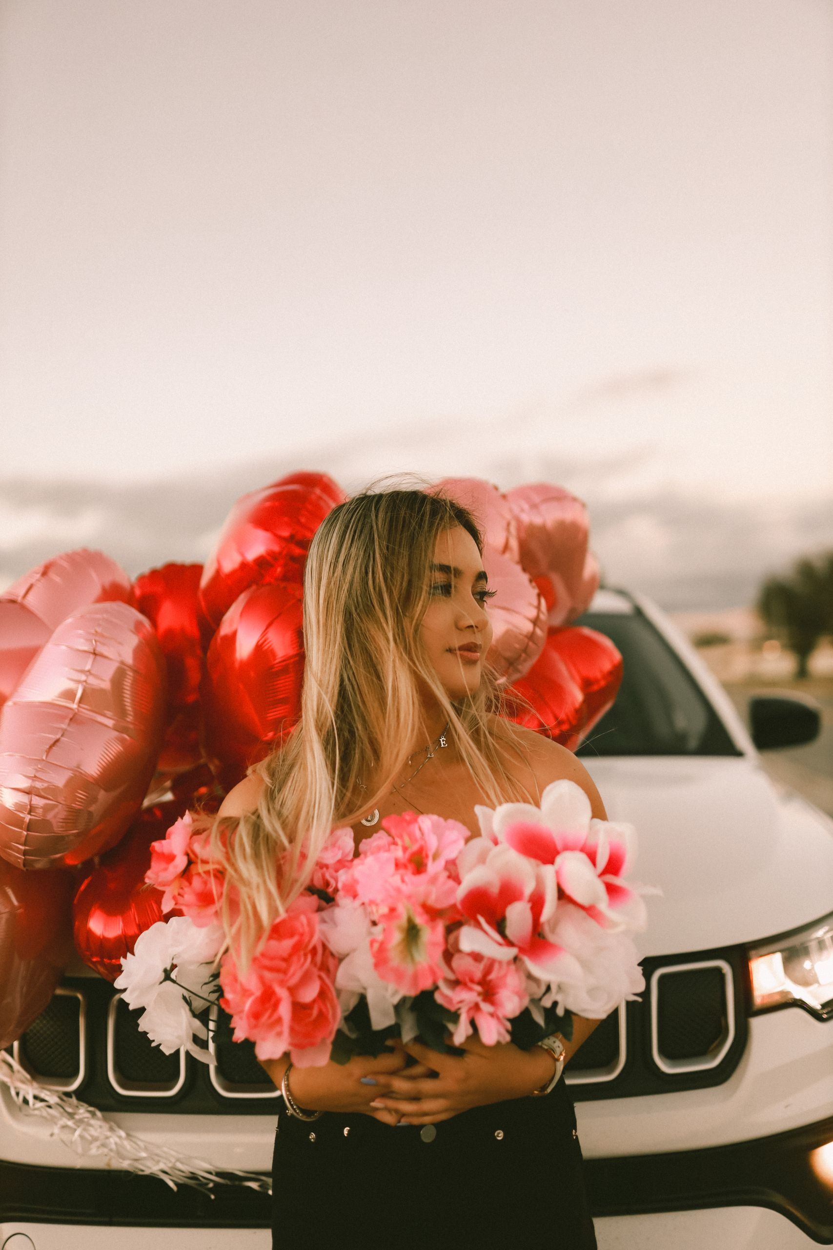 Woman stands by a car holding flowers and balloons, evoking a festive outdoor scene.
