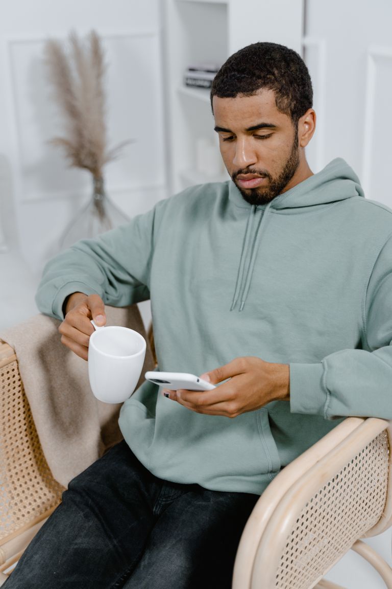 Young man in a hoodie using smartphone and holding coffee mug indoors, experiencing leisure time.
