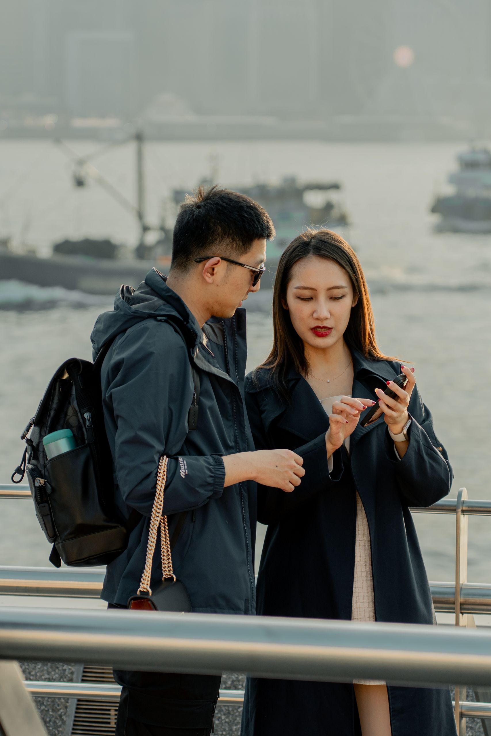 Young urban couple exploring smartphone content on a waterfront pier at sunset.