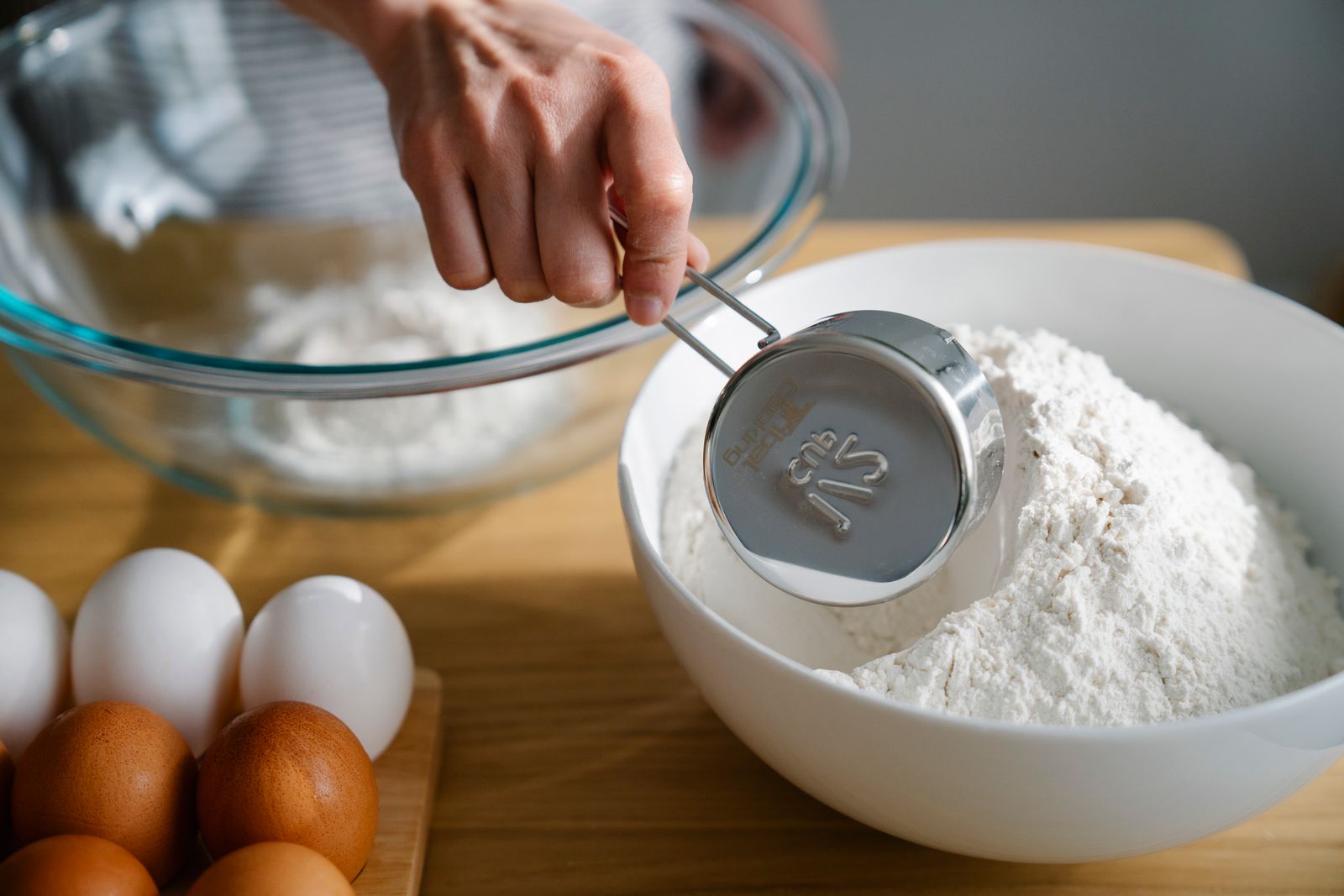 Close-up of a hand measuring flour in a bowl, ready for baking with eggs nearby on the kitchen counter.