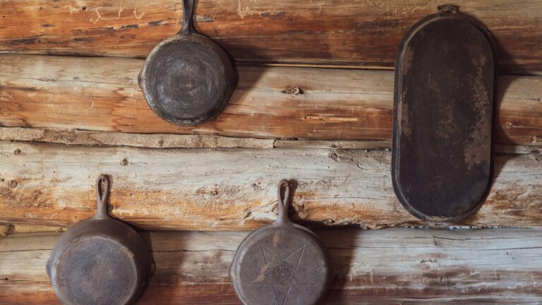 Unsplash photo by Dan Meyers showing three old cast iron skillets hanging on a wall in a pioneer cabin