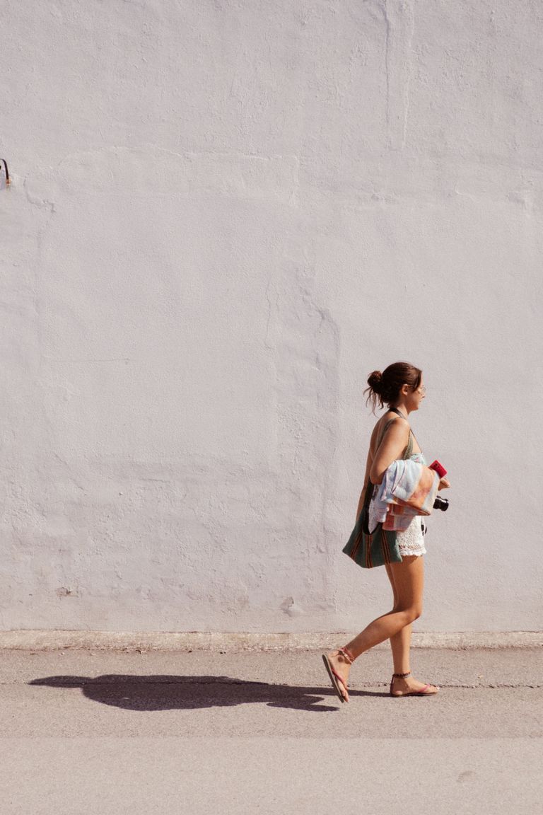 Sunlit scene of a woman walking past a white wall holding various items, casting a shadow.